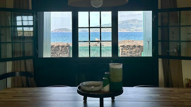 A room with a view of the sea through large windows, a dining table with a bowl and a glass, and a hanging light fixture.
