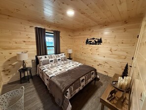 A wooden cabin bedroom with a bed, two bedside tables, a lamp, a chair, and a small table with candles. - New Mountain Cabin, dark skies, and great views! (West Branch Township)