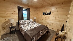 Quarto de cabana de madeira com uma cama, duas mesas de cabeceira, uma cadeira, uma janela com cortinas e um adorno na parede.