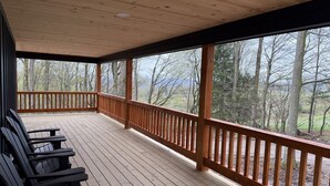 Um deck de madeira coberto com guarda-corpo, cadeiras de madeira e vista para árvores e montanhas ao fundo.