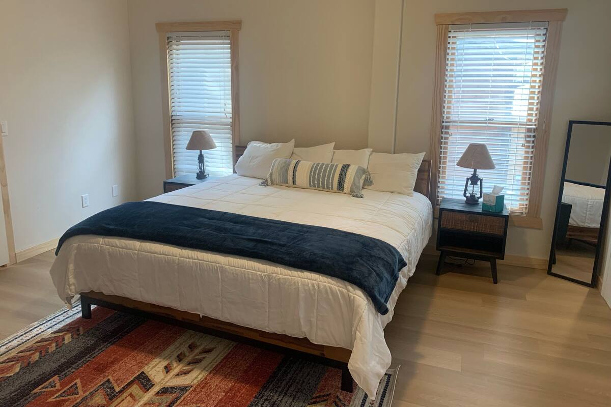 A bedroom with a bed, two bedside tables, a nightstand, a mirror, and two windows with blinds.
