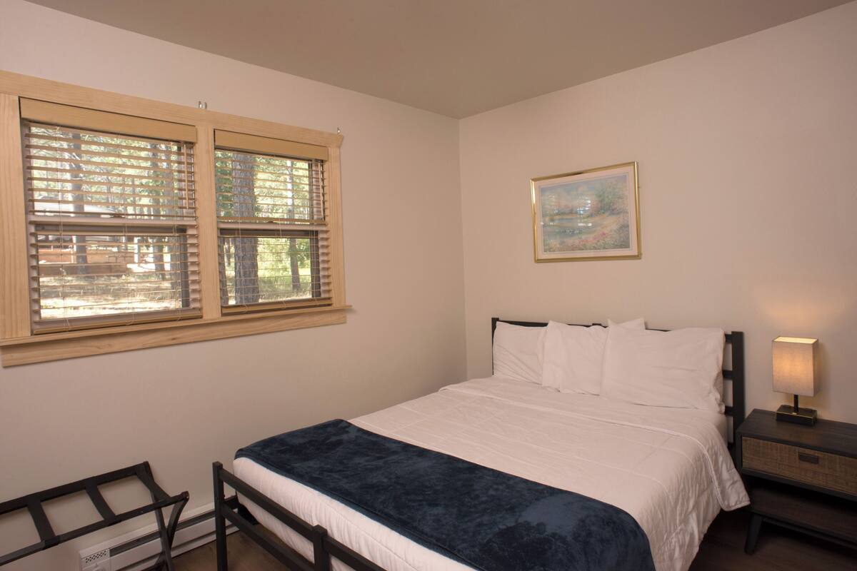 A bedroom with a bed, a nightstand, a window with blinds, and a painting on the wall.