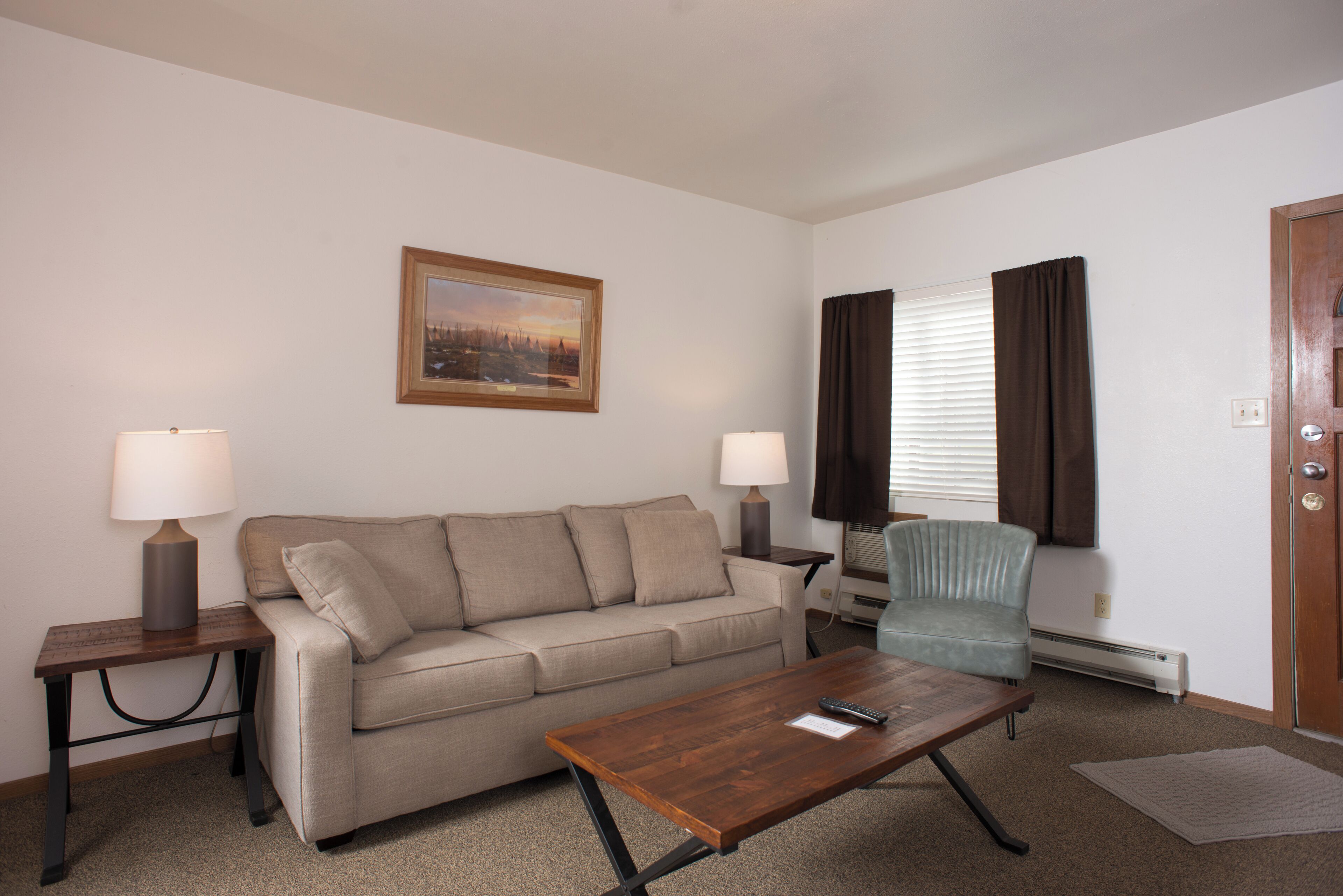 A living room with a beige sofa, two side tables with lamps, a wooden coffee table, a blue armchair, a painting on the wall, and a door with a window.