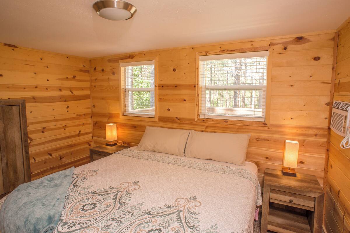 A wooden cabin room with two windows, a bed with a patterned blanket, and bedside lamps.