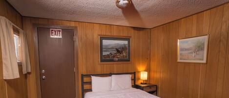 A bedroom with a wooden door marked "EXIT," a bed with white bedding, a nightstand with a lamp, and two framed pictures on the wall.