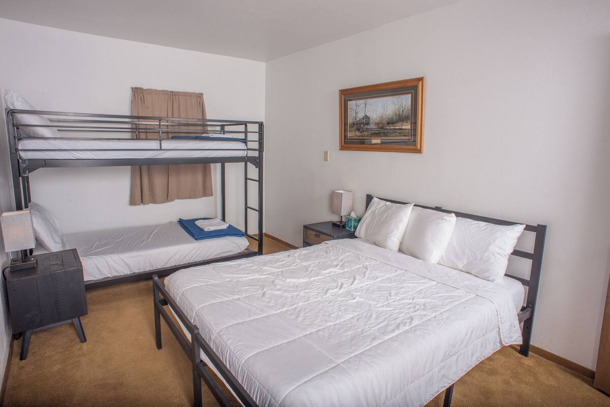 A room with a bunk bed, a single bed, a nightstand, a lamp, and a framed picture on the wall.