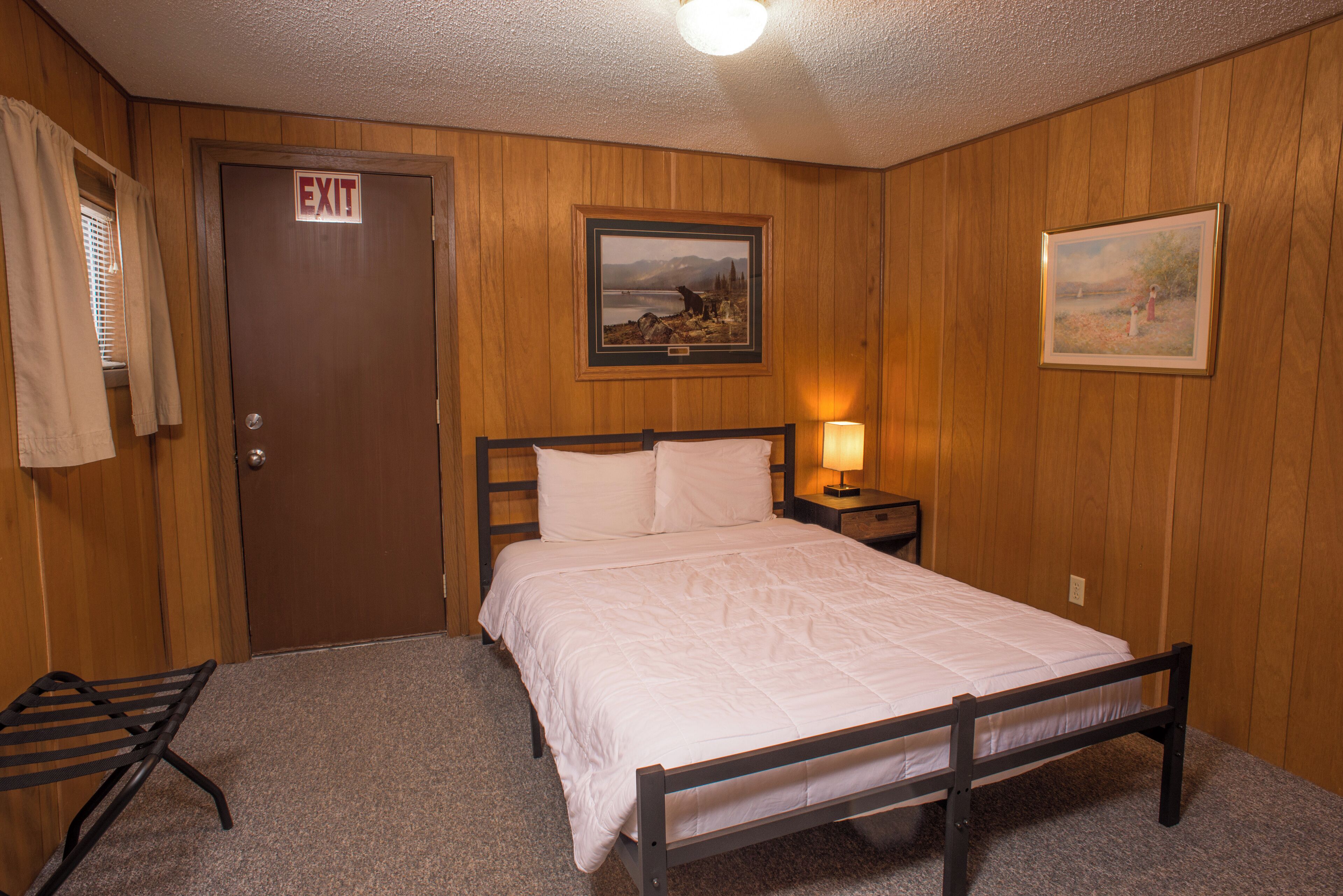 A bedroom with a bed, an exit door, a framed picture, and a nightstand with a lamp.