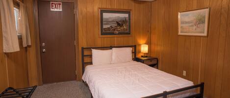 A bedroom with a bed, an exit door, a framed picture, and a nightstand with a lamp.