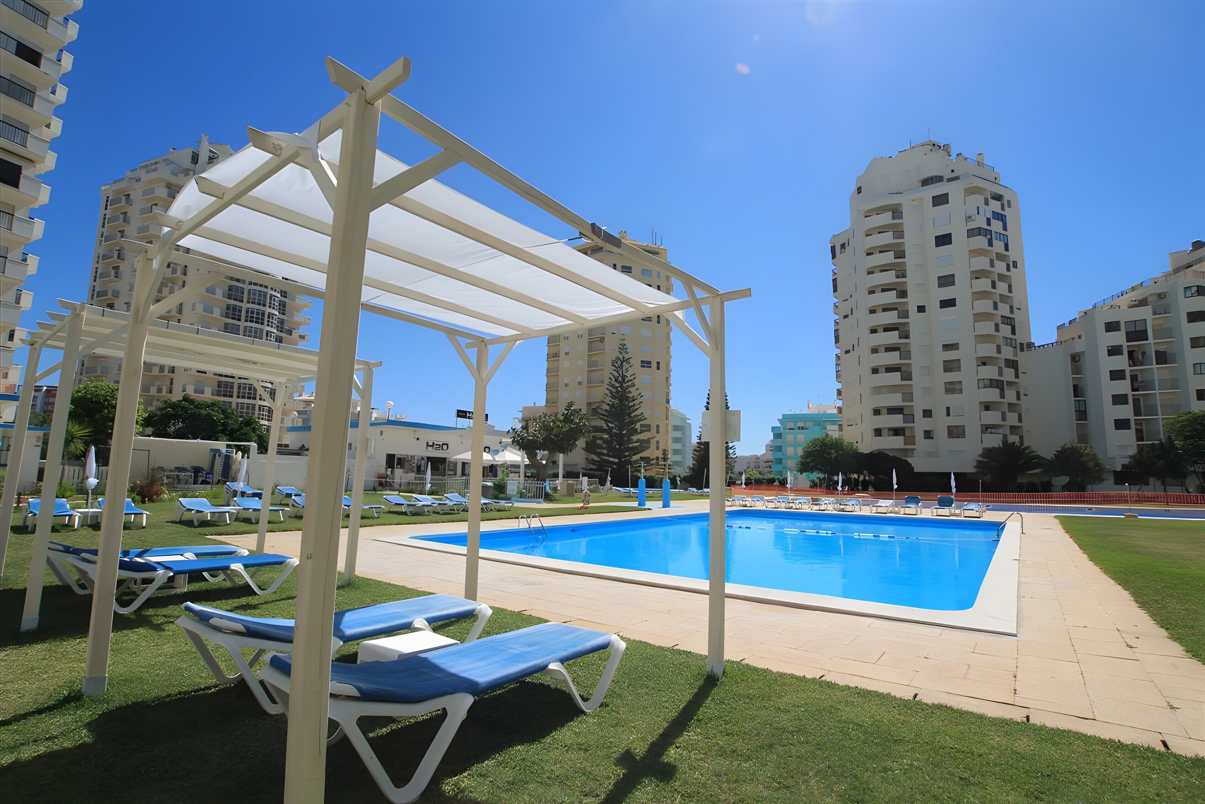 Una piscina con sillones de descanso, una pérgola y edificios altos al fondo.