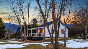 A cabin with a porch, surrounded by trees and snow patches, set against a mountainous backdrop. - Serene Fairview Escape (Fairview)