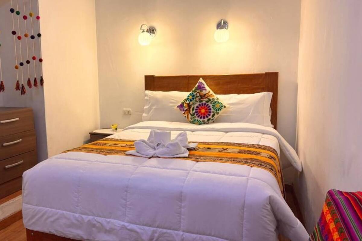 A neatly made bed with a colorful pillow, a wooden nightstand, a wall-mounted light fixture, and a wall decoration.