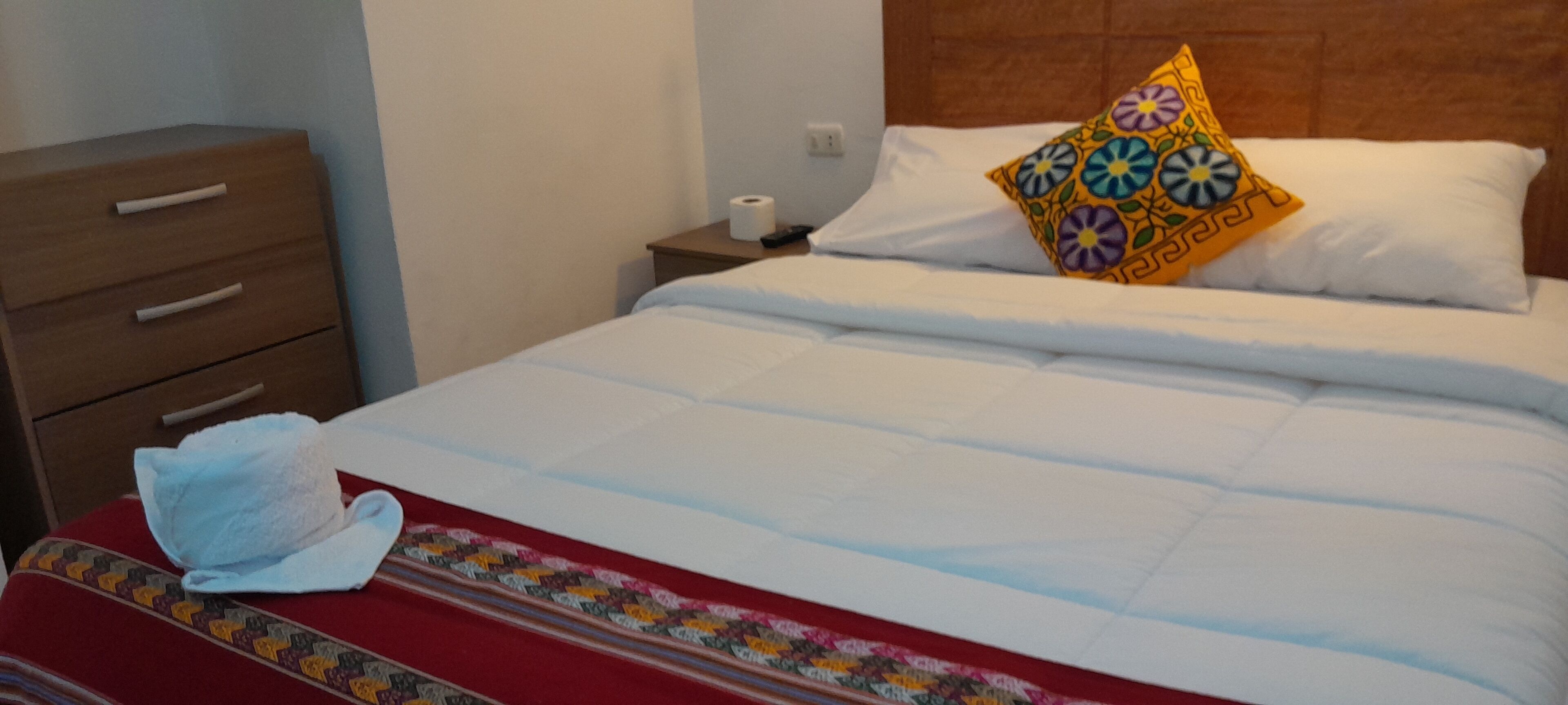 Uma cama bem arrumada com travesseiro estampado, manta listrada em vermelho e branco, e uma toalha branca sobre uma cômoda de madeira.