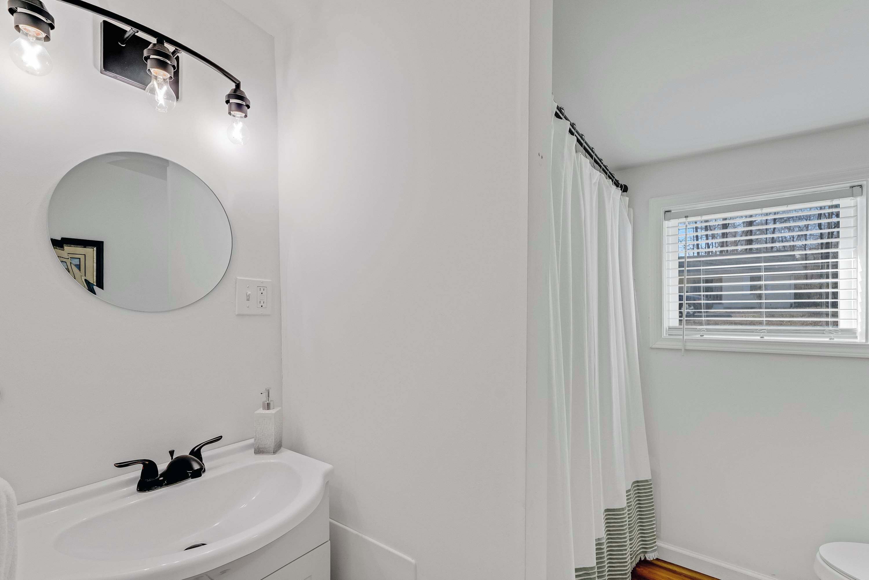 Une salle de bain avec un lavabo blanc, un miroir rond et une fenêtre avec des stores.