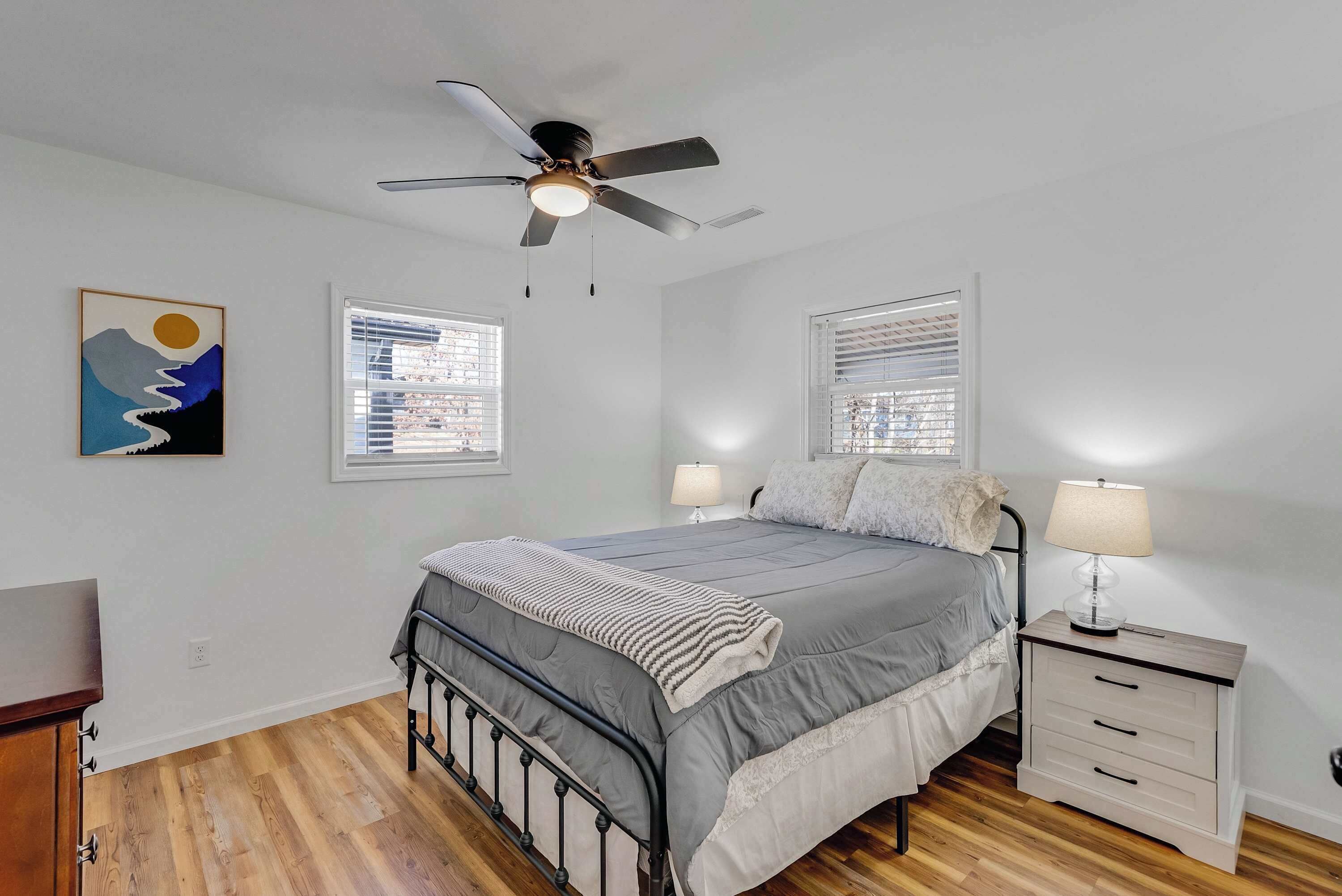 Un dormitorio con cama, mesitas de noche, ventilador de techo y dos ventanas con persianas.