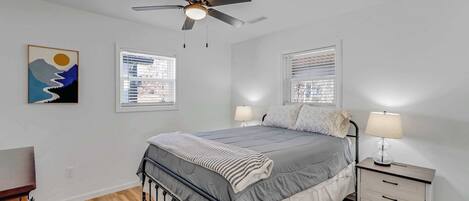 Un dormitorio con cama, mesitas de noche, ventilador de techo y dos ventanas con persianas.