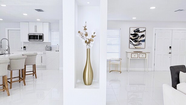 Un espai de vida modern i minimalista amb un columnà central blanc, un vas daurat amb flors seques, un sofà blanc, un armari negre, una taula de consol·la blanca i una pintura abstracta en blanc i negre.