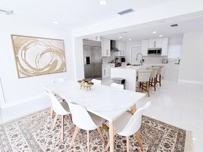 Una cocina moderna con armarios blancos, una isla central y un comedor con una mesa y sillas blancas.