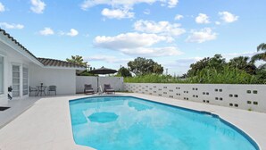 A swimming pool with a white tiled deck, a covered patio area with chairs, and a white building with a tiled roof. - Charming 3BR/2BA Pool Home in Sarasota, Total Comfort, Minutes to Siesta Key! (Sarasota)