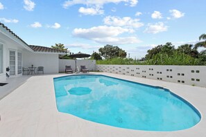 Ein Swimmingpool mit weiß gefliestem Deck, eine überdachte Terrasse mit Stühlen und ein weißes Gebäude mit einem gefliesten Dach.