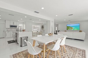 A modern kitchen with white cabinetry, a dining area with a white table and chairs, and a living room with a sofa and a TV displaying a scenic view. - Charming 3BR/2BA Pool Home in Sarasota, Total Comfort, Minutes to Siesta Key! (Sarasota)