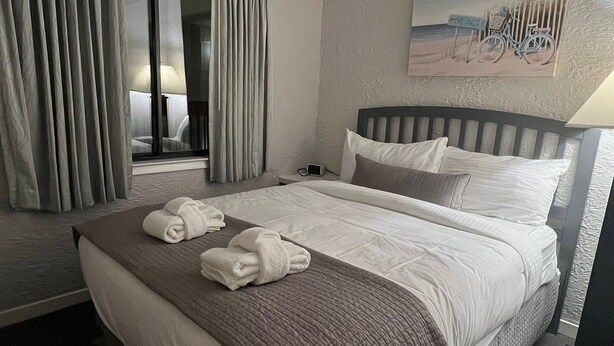 Une chambre d’hôtel avec un lit, une fenêtre avec des rideaux, un tableau au mur et une lampe.