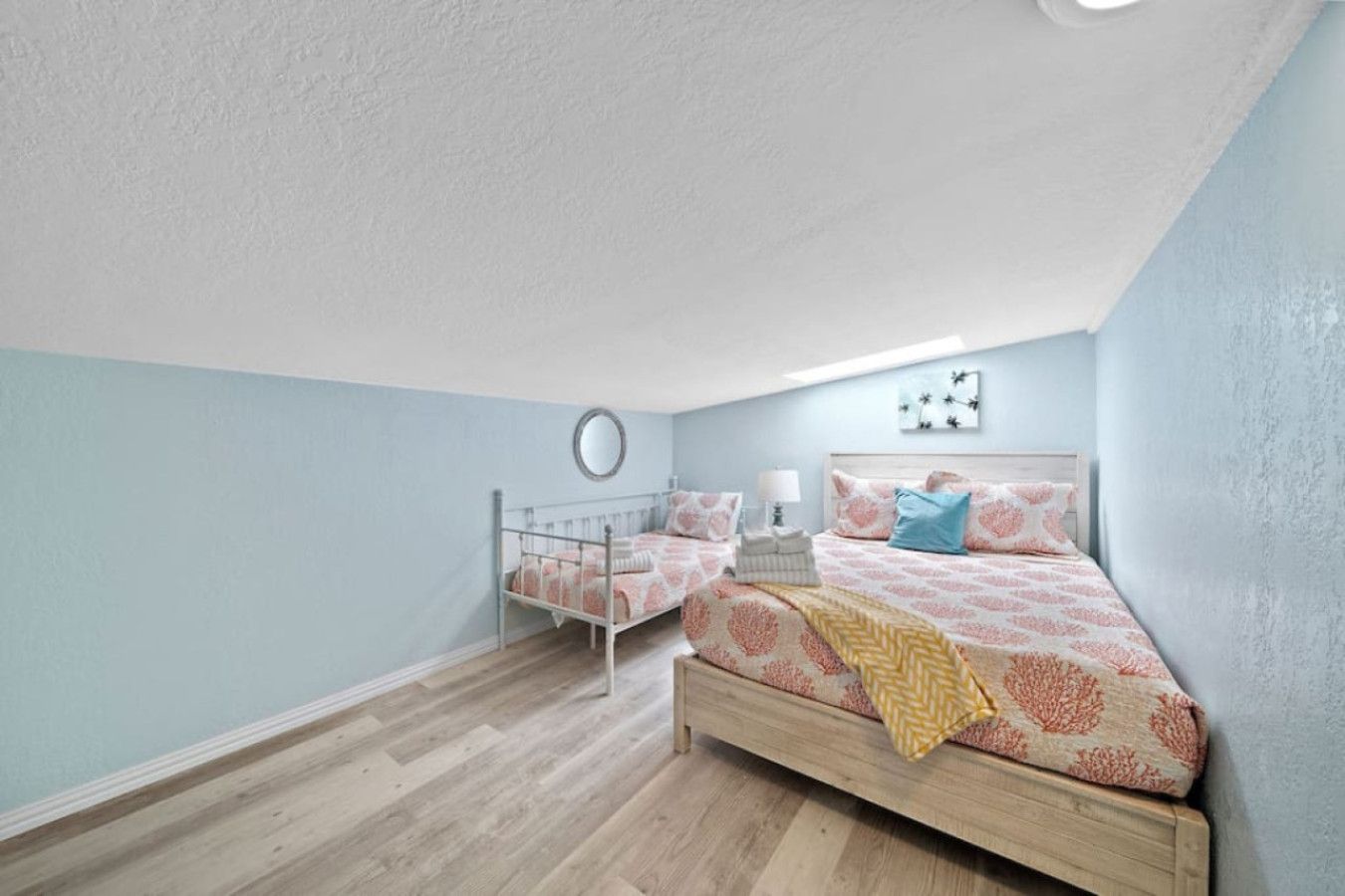 A bedroom with a single bed, a smaller bed, a bedside table with a lamp, and a wall-mounted mirror.