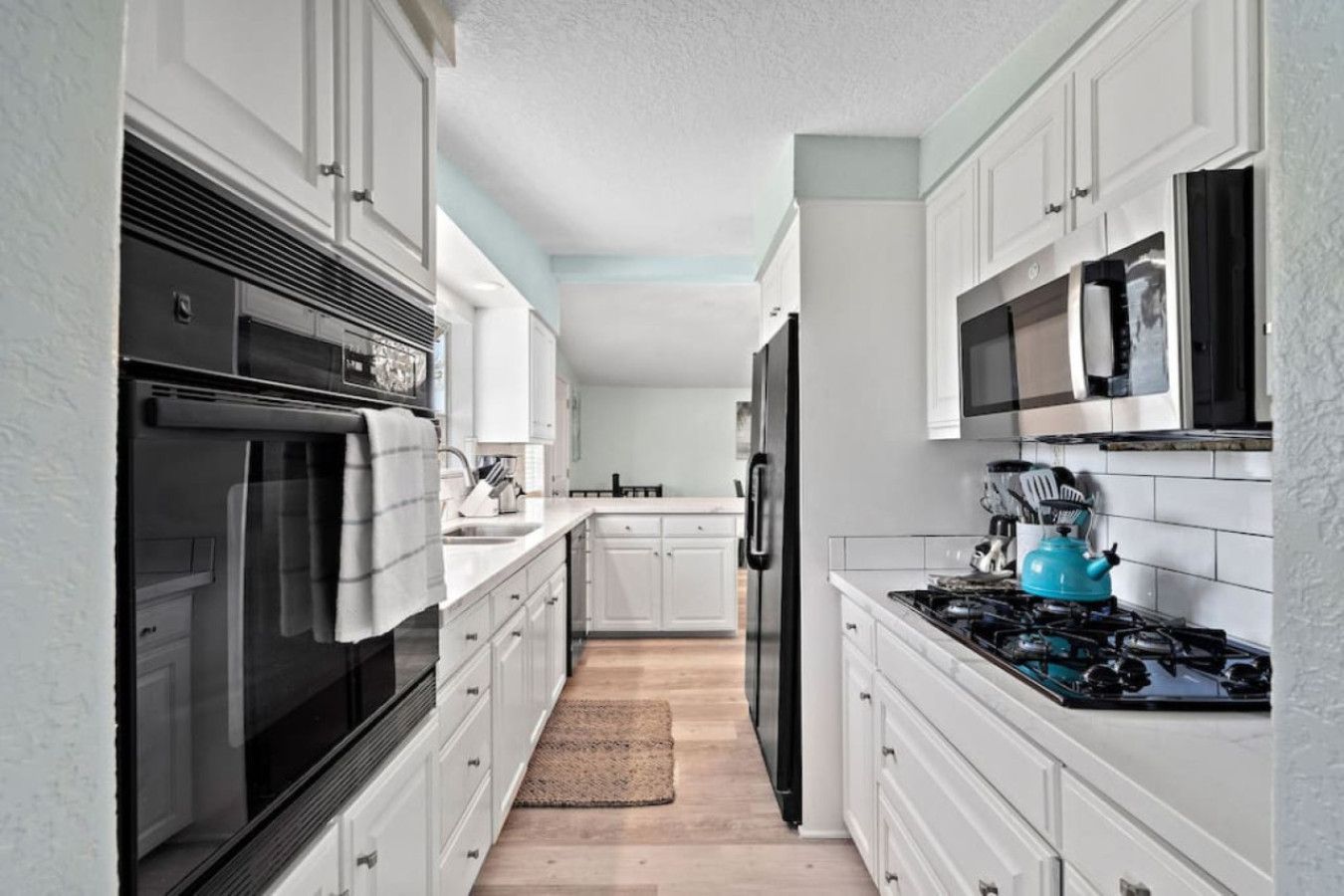 A narrow kitchen with white cabinets, a black oven, a blue kettle, and a microwave.