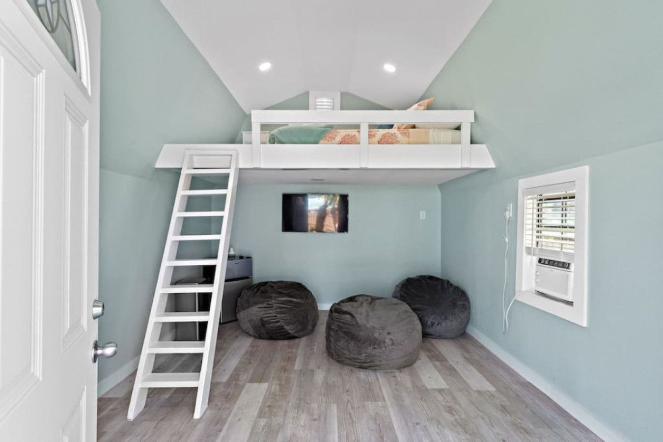 A room with a loft bed, a ladder, two bean bags, a small TV, and a window with blinds.
