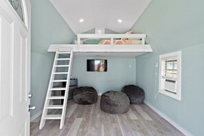 Una habitación con una cama alta, una escalera, dos puffs, un televisor pequeño y una ventana con persianas.
