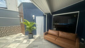 En overdækket terrasse med en brun lædersofa, en potteplante samt udsigt til et swimmingpool og et udendørs spiseområde.