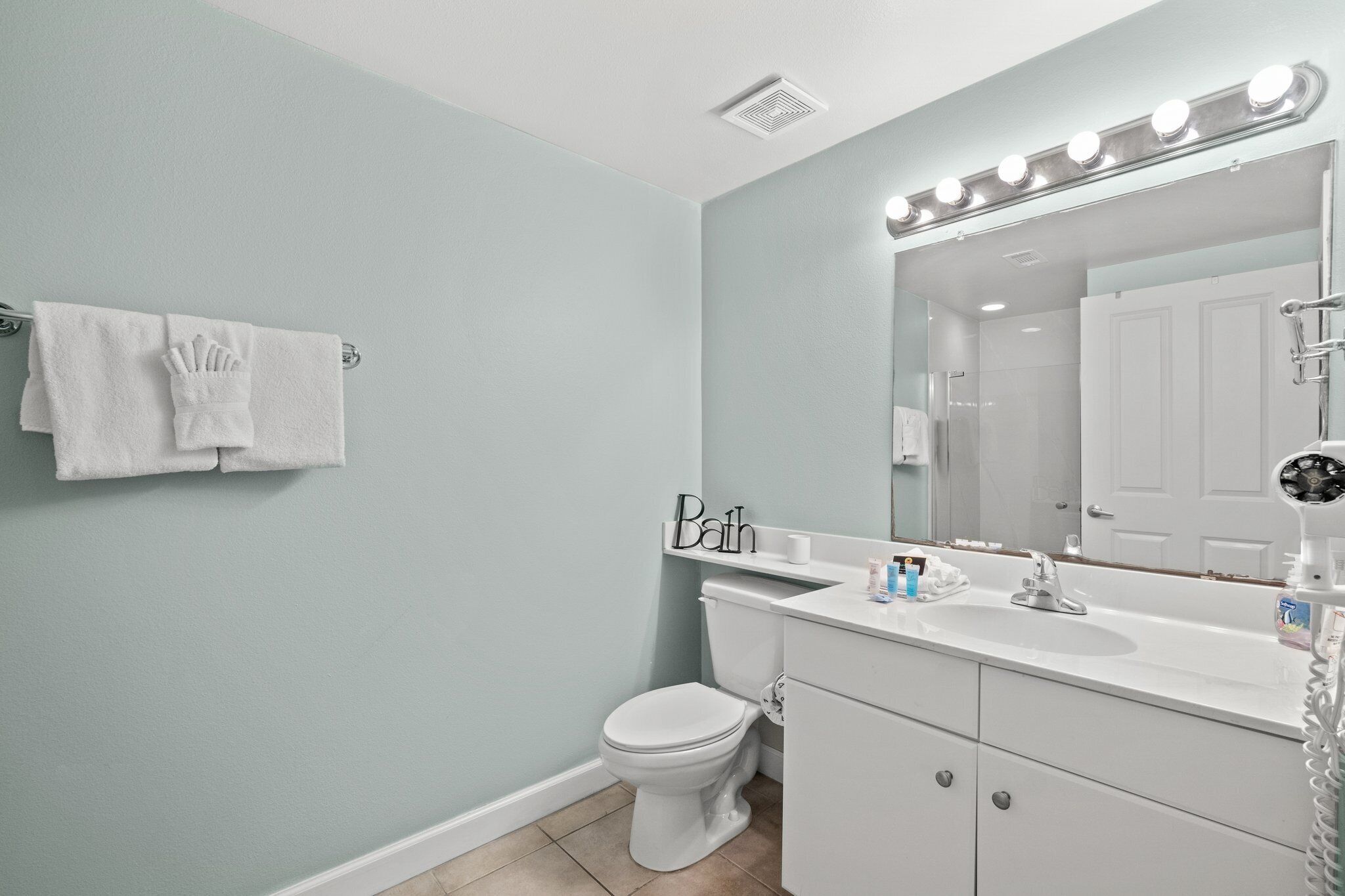 Une salle de bain avec un lavabo blanc, un miroir et des toilettes. On y trouve un porte-serviettes avec une serviette et une pancarte indiquant « Salle de bain ».