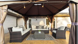 A covered patio area with wicker furniture and a brick wall backdrop. - Resort Style 4BR Plano Retreat - Pool, Spa & Entertainment (Plano)