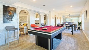 A pool table in a modern living room with a brick wall, a sofa, and a dining area in the background. - Resort Style 4BR Plano Retreat - Pool, Spa & Entertainment (Plano)