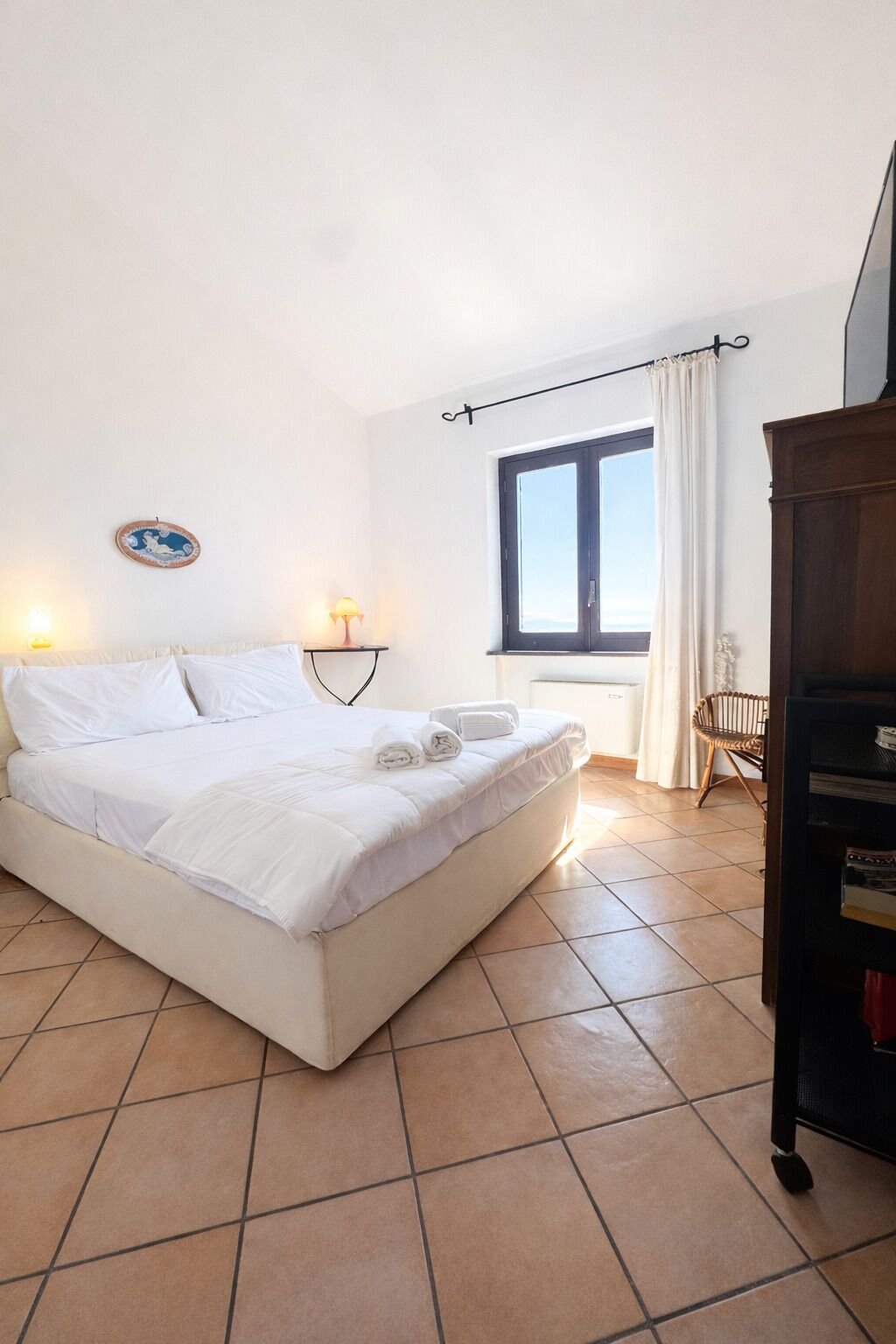 Un dormitorio con cama, lámparas de noche, una ventana con cortinas, un radiador y un armario de madera.