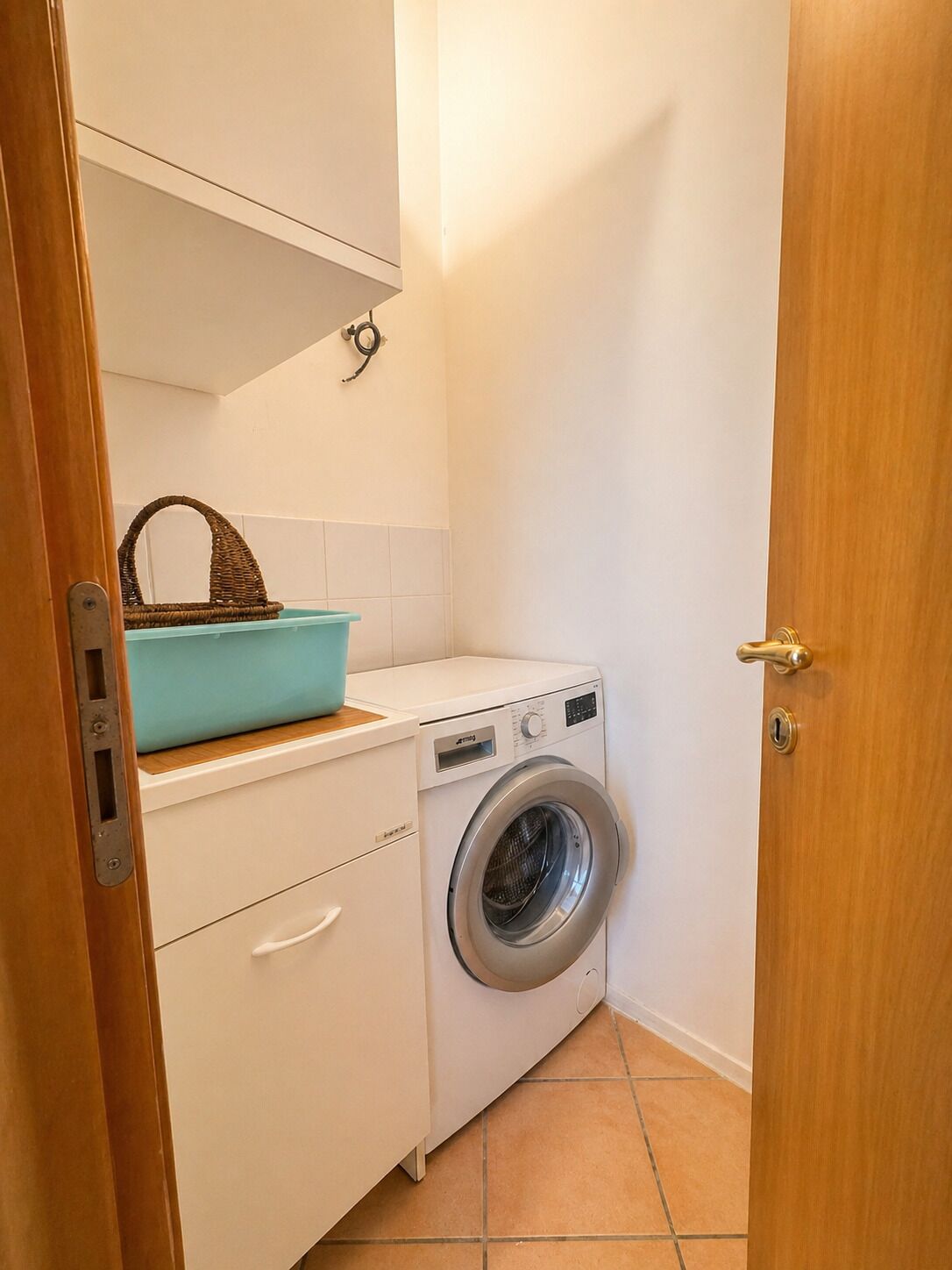 Un lavadero pequeño con lavadora, tendedero y cesto sobre un mueble.