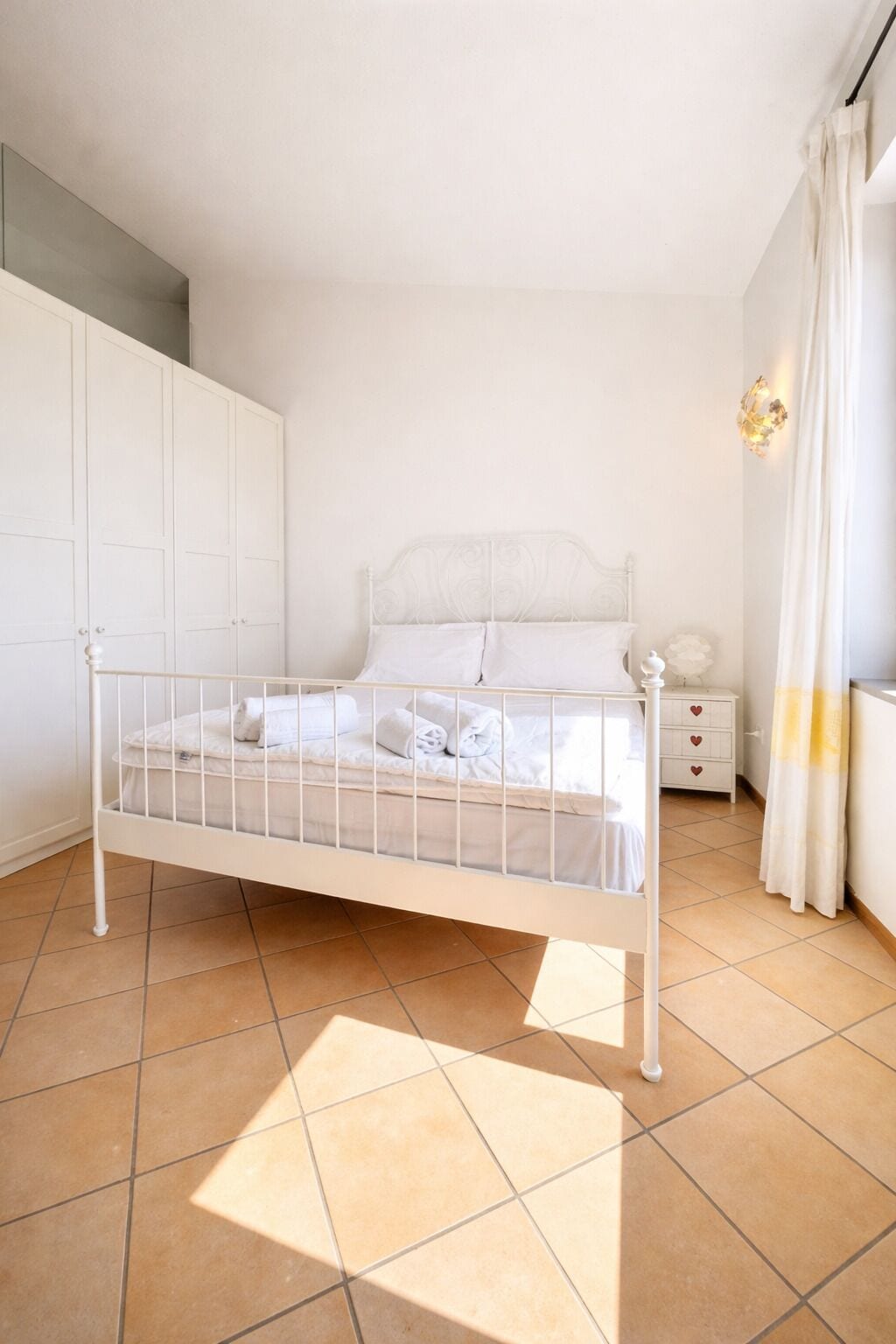 A white metal bed in a room with tiled flooring, a wardrobe, a bedside table with a lamp, and a curtain.
