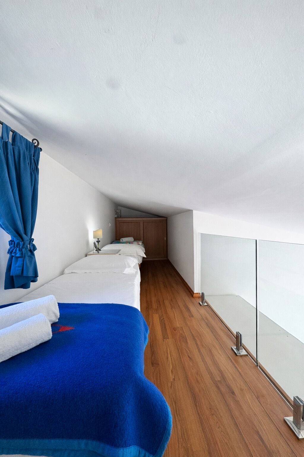 Een hotelkamer met een houten vloer, een bed met een blauwe deken, een blauw gordijn en een klein nachtkastje met een lamp.