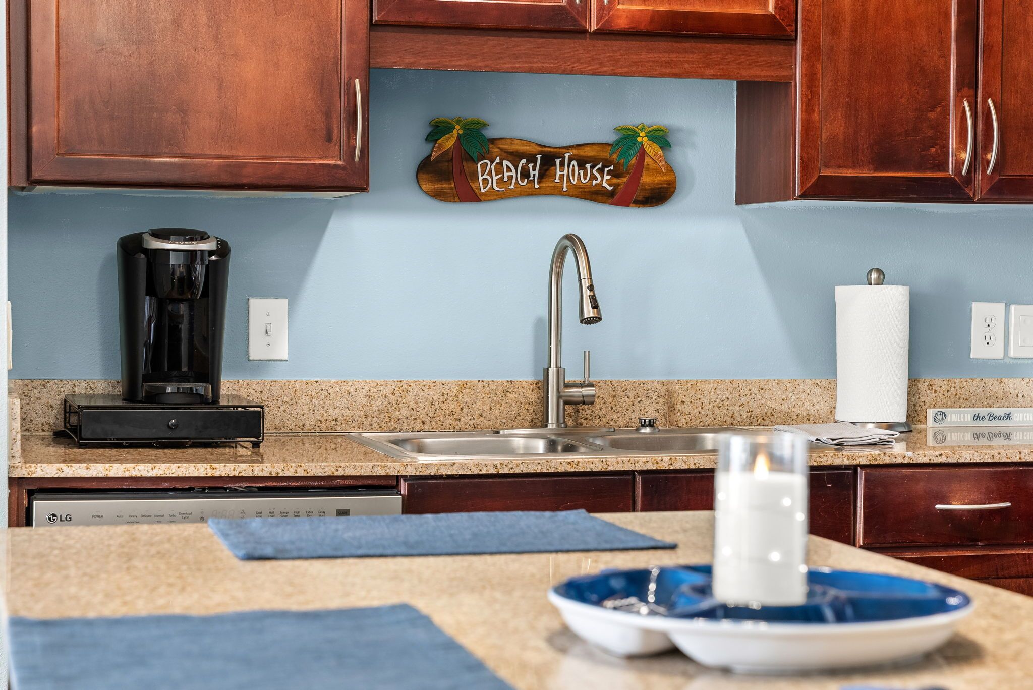 A kitchen with wooden cabinets, a coffee maker, a sink, and a decorative sign that reads "Beach House".