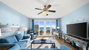 A living room with a large TV, blue sofas, a coffee table, and a balcony with a view of the ocean. - Dolphin Views! Laketown Wharf - 7th Floor! (Panama City Beach)