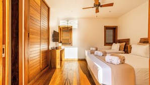 Une chambre d’hôtel avec un plancher en bois, un lit avec du linge de lit blanc, une tête de lit en bois, un ventilateur de plafond et une fenêtre avec des volets en bois.