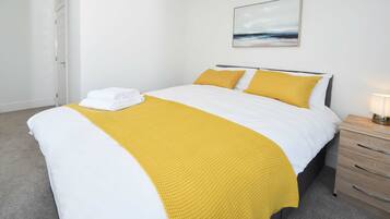 A neatly made bed with white and yellow bedding, a single yellow throw blanket, and a folded white towel on the foot of the bed.