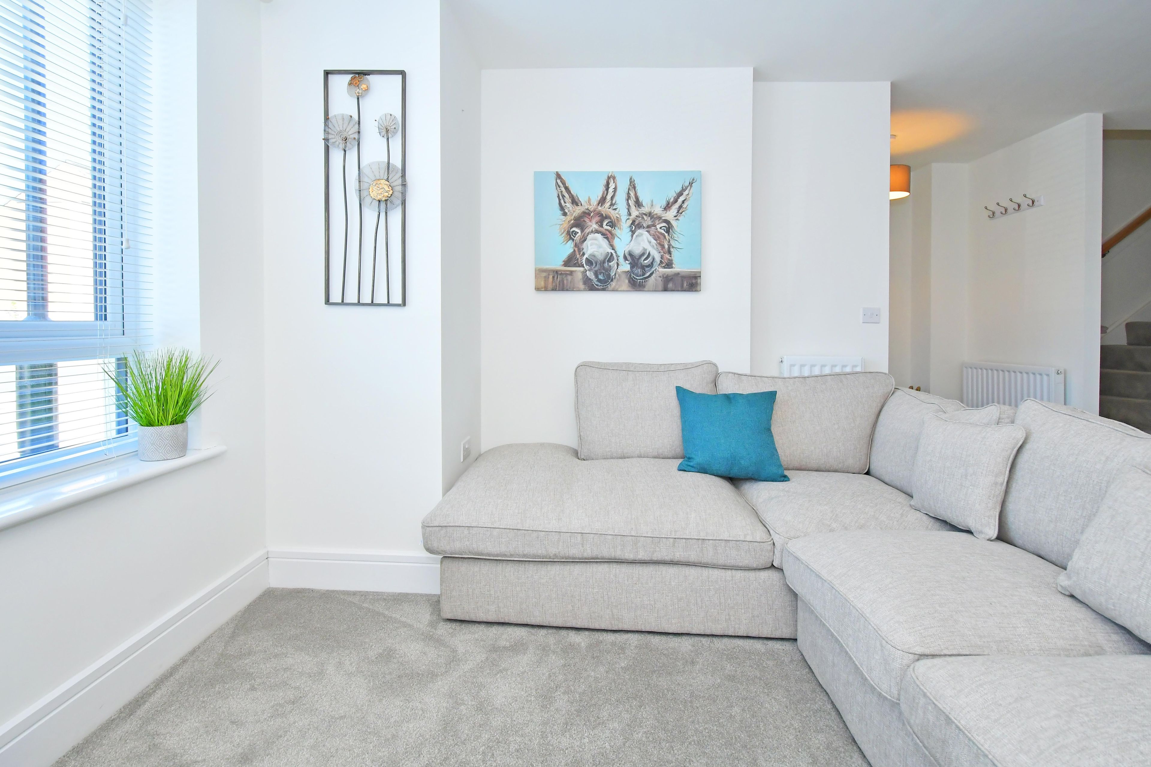 A living room with a grey sectional sofa, a blue pillow, a framed artwork of donkeys, and a potted plant on a windowsill.