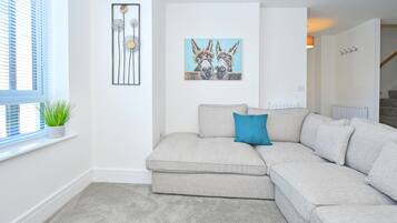 A living room with a grey sectional sofa, a blue pillow, a framed artwork of donkeys, and a potted plant on a windowsill.