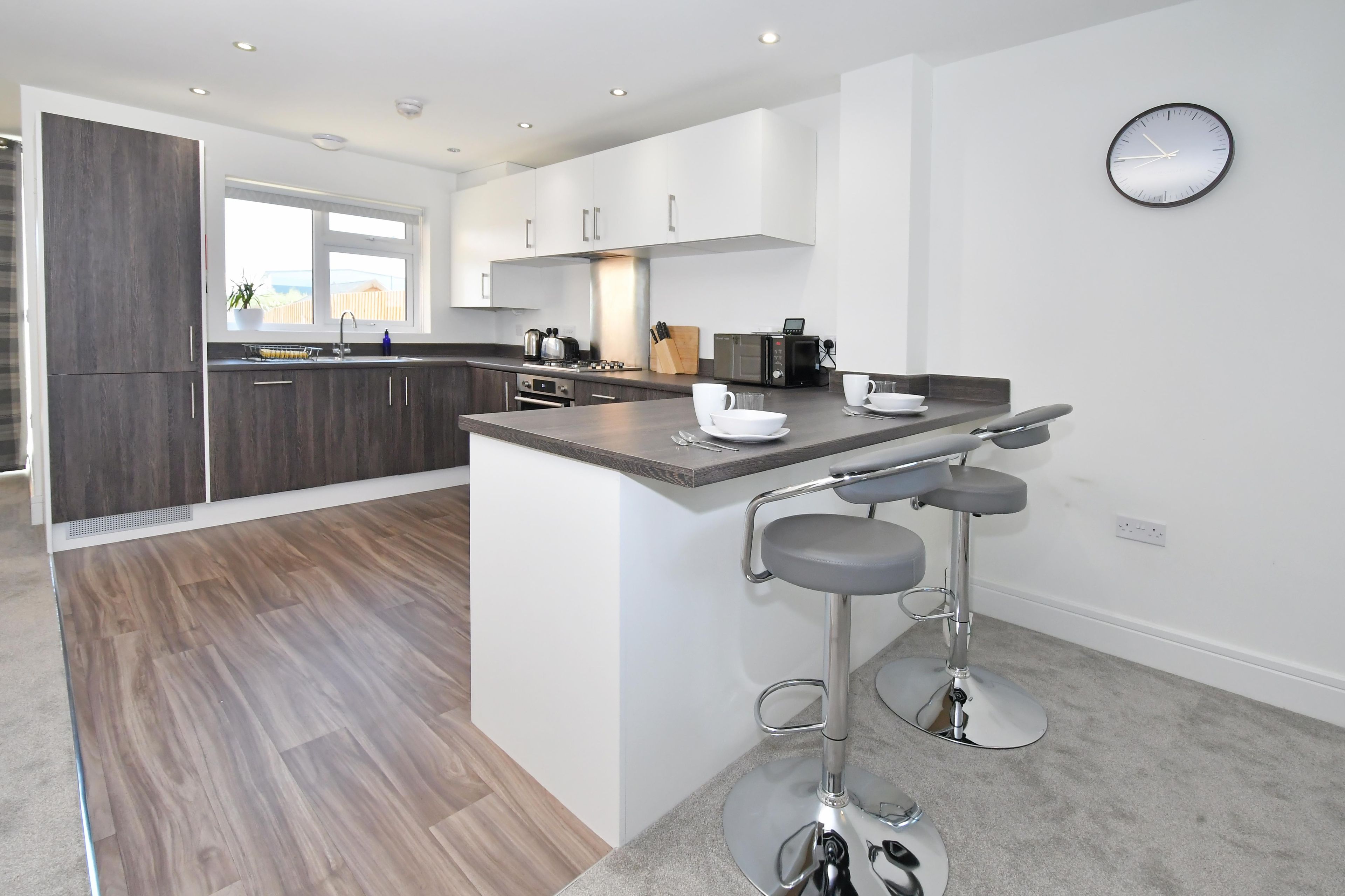A modern kitchen with a central island, bar stools, and a wall-mounted clock.