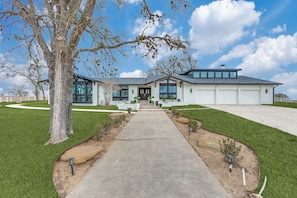 Une maison moderne à deux étages, avec une allée en béton menant à la porte d’entrée, entourée d’une pelouse bien entretenue et d’un grand arbre.