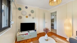 A living room with a flat-screen TV, a white coffee table, a wicker lamp, and a wall decorated with plates. - Lisbon Nearby. Surrounded by Nature. Beaches Always Close. (Trafaria)