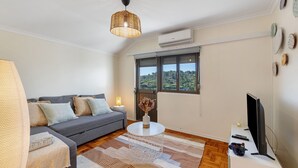 A living room with a sofa, a coffee table, a TV, and a window with a view. - Lisbon Nearby. Surrounded by Nature. Beaches Always Close. (Trafaria)