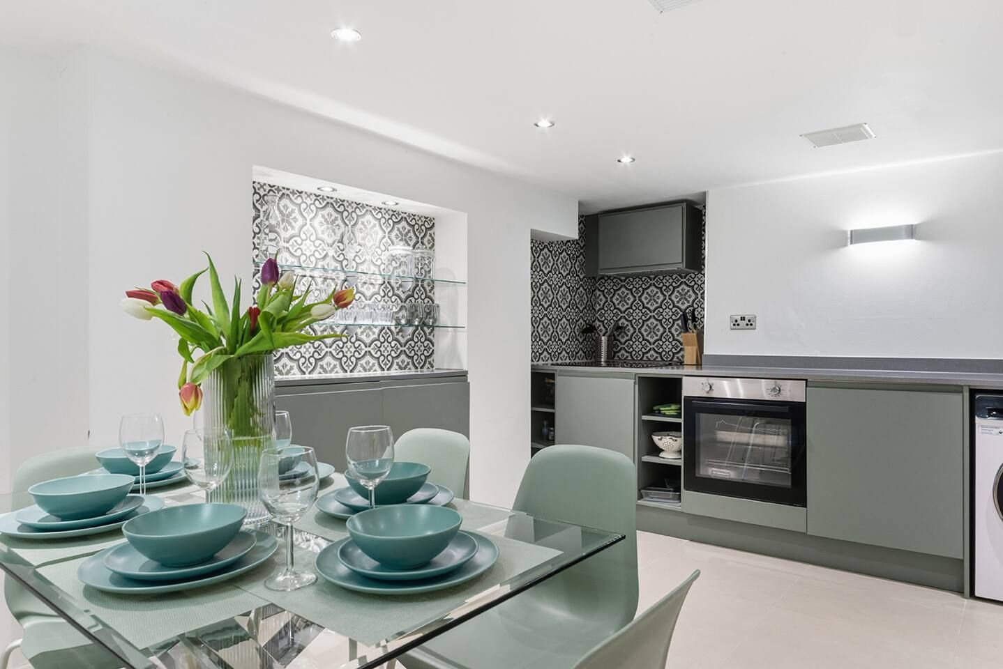 A modern kitchen with a dining area, featuring a glass table, teal dinnerware, and a vase with flowers.