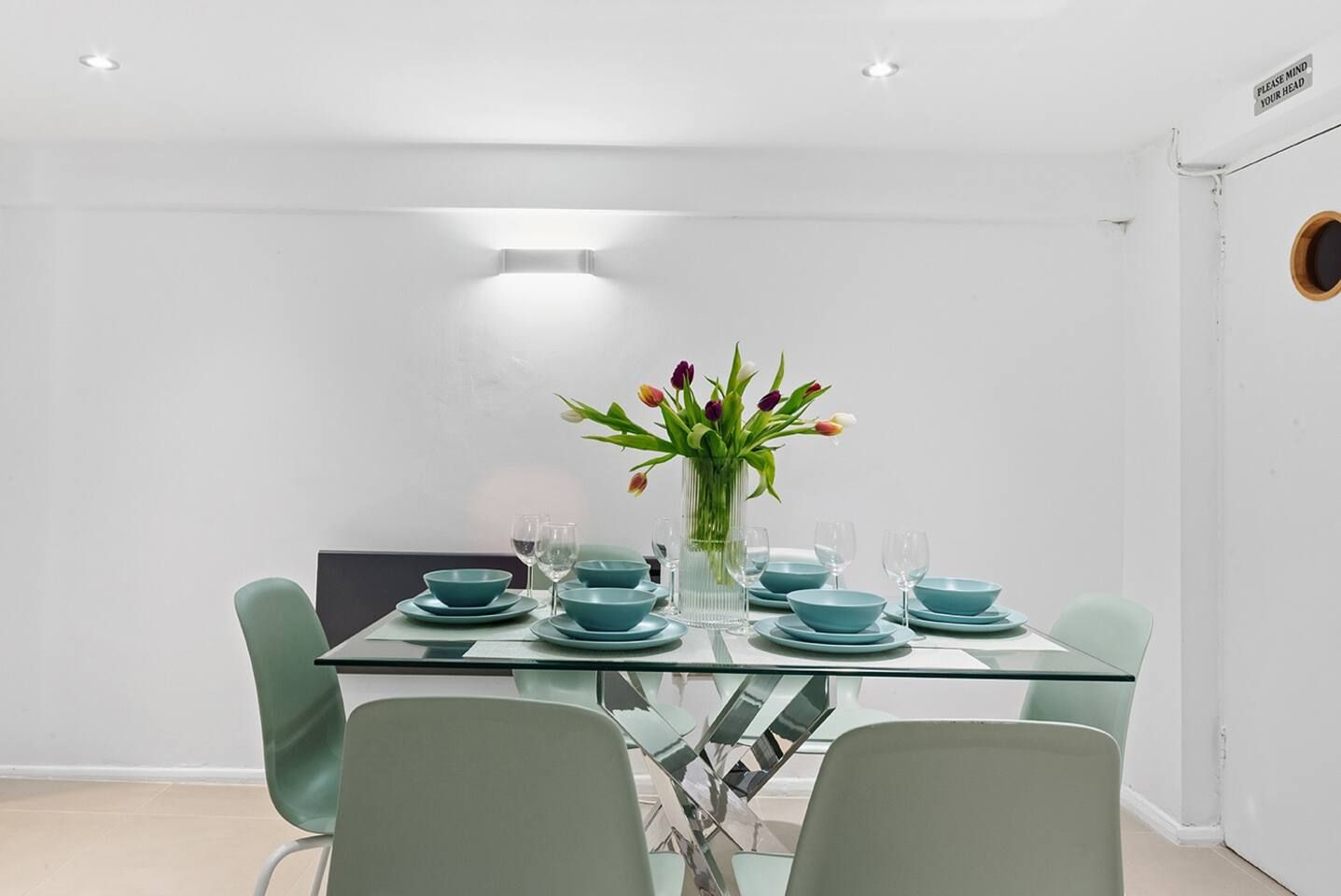 A dining area with a glass table set for four, surrounded by four chairs, and a vase with flowers on the table.