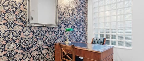 Une pièce avec un papier peint à motifs, un bureau avec une chaise, un miroir et une fenêtre avec des stores.